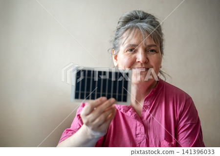 Smiling woman holding a portable solar panel indoors with soft lighting. Renewable energy green technology and solar energy education Smiling woman holding a portable solar panel indoors with soft lighting. Renewable energy green technology and solar energy education 141396033
