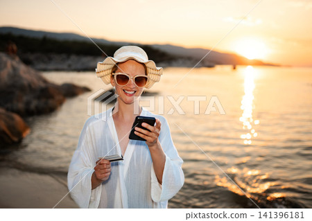 Woman holding smartphone and credit card on beach at sunset Woman holding smartphone and credit card on beach at sunset 141396181