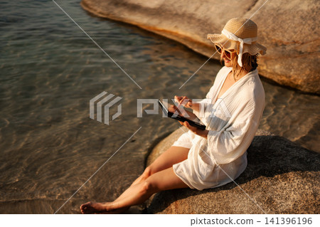 Woman freelancer working on tablet by ocean under sunshine Woman freelancer working on tablet by ocean under sunshine 141396196