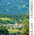 Panoramic view of the countryside beneath the Julian Alps in Slovenia, featuring a picturesque village with a church at its center surrounded by green fields and forests. 141396603