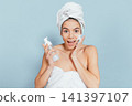 Happy woman in towel, using skincare product against blue backdrop 141397107