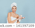 Smiling woman in towel holds spray bottle against light blue background 141397109