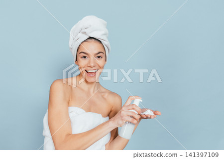 Smiling woman in towel holds spray bottle against light blue background Smiling woman in towel holds spray bottle against light blue background 141397109