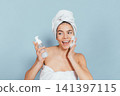 Happy woman wraps towel around head while applying face wash 141397115