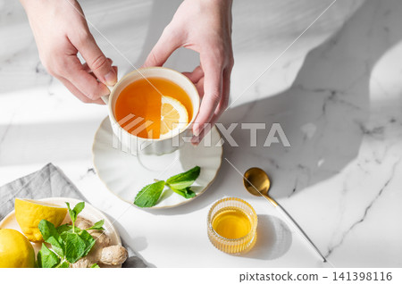 Hand holding a cup of tea with lemon, ginger and mint on a marble background Hand holding a cup of tea with lemon, ginger and mint on a marble background 141398116