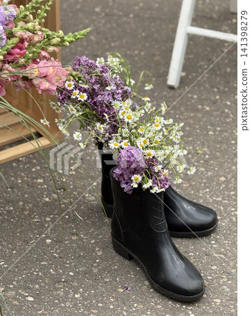 Flowers arranged in black boots creating unique garden decoration outdoors near wooden structure Flowers arranged in black boots creating unique garden decoration outdoors near wooden structure 141398279