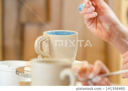 A woman steadily glazes a white ceramic mug on a rotating tray in a pottery studio; A woman steadily glazes a white ceramic mug on a rotating tray in a pottery studio; 141398460