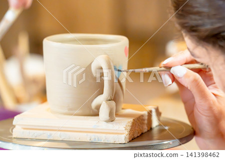 A woman steadily glazes a white ceramic mug on a rotating tray in a pottery studio; A woman steadily glazes a white ceramic mug on a rotating tray in a pottery studio; 141398462