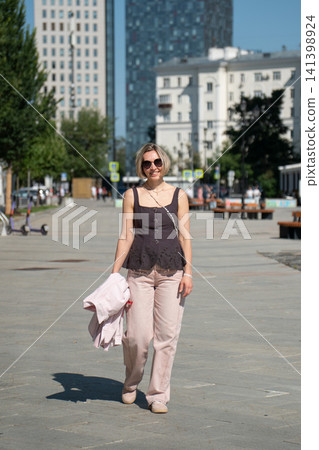 Woman approaches camera during summer walk on Shkolnaya Street in Moscow carrying jacket. Urban lifestyle, city travel, leisure, confidence, exploration, warm weather, freedom 141398924