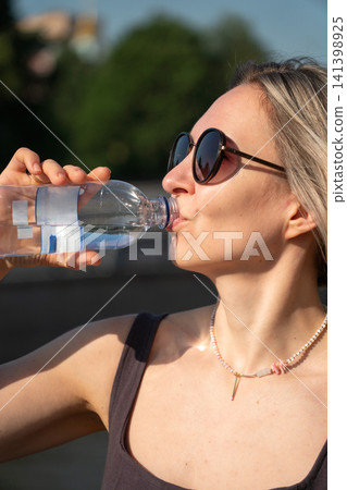 Vertical portrait of woman drinking water outdoors in Moscow during summer. Hydration, wellness, heat relief, healthy habit, active lifestyle, travel, self care, city life Vertical portrait of woman drinking water outdoors in Moscow during summer. Hydration, wellness, heat relief, healthy habit, active lifestyle, travel, self care, city life 141398925