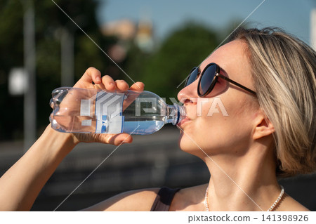 Woman drinking bottled water outdoors in Moscow. Hydration, summer heat, healthy lifestyle, wellbeing, refreshment, self care, fitness, urban living 141398926