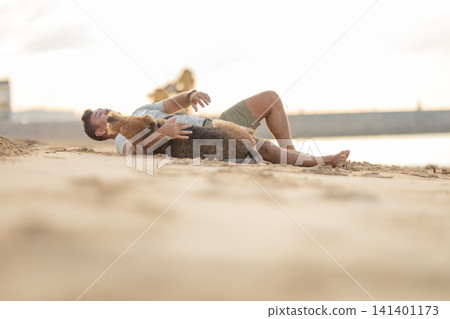 Man lying on sandy beach with dog, enjoying playful moment together, water and distant structures visible in the background during golden hour. Airedale Terrier Man lying on sandy beach with dog, enjoying playful moment together, water and distant structures visible in the background during golden hour. Airedale Terrier 141401173