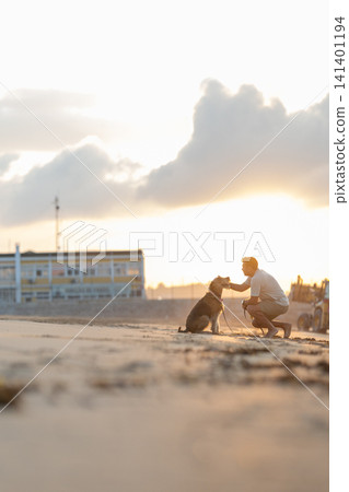 Man kneeling on sandy beach interacts with brown dog during sunset, with distant beach houses and clouds visible in the background. Airedale Terrier Man kneeling on sandy beach interacts with brown dog during sunset, with distant beach houses and clouds visible in the background. Airedale Terrier 141401194