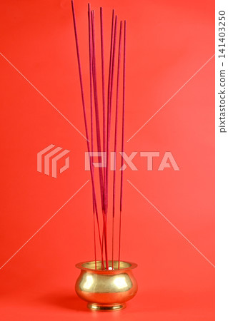 A close-up shot of several magenta incense sticks standing upright in a traditional polished brass bowl, set against a solid vibrant red background 141403250