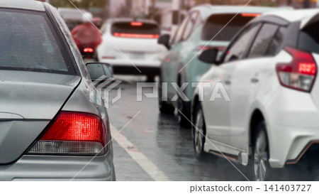 Rear view of multiple cars driving on a wet road during rainy weather. Reflections on the asphalt and red tail lights highlight a slow moving urban traffic situation. 141403727