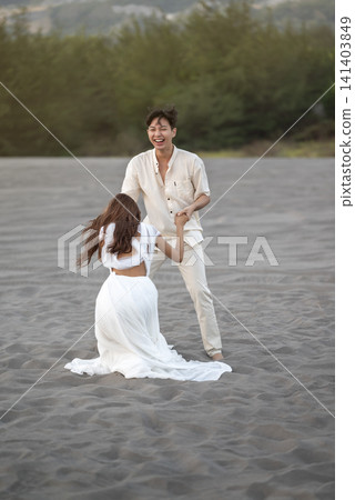 A young Indonesian southeast asian romantic couple in light clothing holding hands and dancing on expansive sand dunes during an outdoor photoshoot in a beautiful landscape A young Indonesian southeast asian romantic couple in light clothing holding hands and dancing on expansive sand dunes during an outdoor photoshoot in a beautiful landscape 141403849
