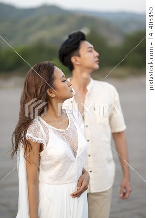 A loving young Indonesian southeast asian couple posing for their pre-wedding photoshoot on a beach. The woman wears a white lace dress while the man stands behind in a light casual shirt A loving young Indonesian southeast asian couple posing for their pre-wedding photoshoot on a beach. The woman wears a white lace dress while the man stands behind in a light casual shirt 141403850