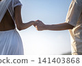 Close-up of a loving couple holding hands outdoors, showing a woman in a white backless dress and a man in a light shirt against a clear sky 141403868