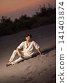 A young Indonesian southeast asian man in a stylish white linen shirt and pants sits relaxed on a sand dune, posing under a dramatic sunset sky at dusk 141403874