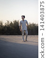 A handsome young Indonesian southeast asian man stands confidently on a sandy dune, wearing a casual linen shirt and trousers, backlit by the warm golden light of the sun 141403875