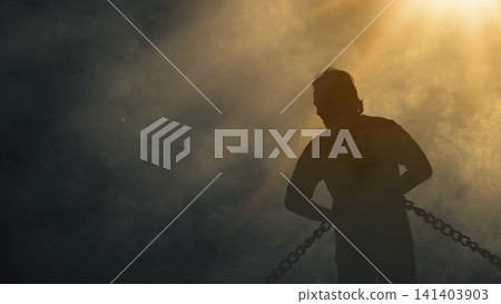 Dramatic silhouette of a man bound in heavy chains, standing under a bright golden light beam illuminating a dark, smoky sky, symbolizing struggle, captivity, and hope of freedom 141403903