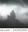 A conceptual silhouette of a man sitting bound in heavy chains on a rocky hill under a dark, stormy, and misty sky, symbolizing depression, addiction, captivity, and mental struggles 141403905