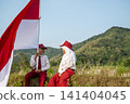 Two Indonesian school children sitting in a sunny field holding the national flag of Indonesia, celebrating patriotism, freedom. Hari Kemerdekaan, Merdeka, and Dirgahayu Indonesia on 17 Agustus 141404045