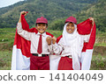 Two Indonesian elementary school students in uniform smiling and holding the red and white national flag in a rural green landscape, celebrating independence day. Hari Kemerdekaan Dirgahayu Indonesia 141404049
