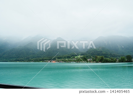Scenic Cruise View of Lake Brienz from a Passenger Boat with Swiss Chalets Village, Alpine Mountains, Turquoise Lake and Mist, Switzerland. Picturesque Landscape and Famous Europe Travel Destination Scenic Cruise View of Lake Brienz from a Passenger Boat with Swiss Chalets Village, Alpine Mountains, Turquoise Lake and Mist, Switzerland. Picturesque Landscape and Famous Europe Travel Destination 141405465