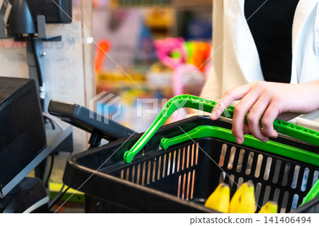 A Close-Up of a Person's Hand Placing Green Shopping Baskets at a Checkout Counter Surrounded by Colorful Toys and Grocery Items. A Close-Up of a Person's Hand Placing Green Shopping Baskets at a Checkout Counter Surrounded by Colorful Toys and Grocery Items. 141406494