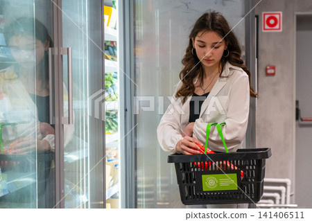 A Young Woman Selects Fresh Produce from a Supermarket Freezer While Carrying a Shopping Basket Filled with Groceries. A Young Woman Selects Fresh Produce from a Supermarket Freezer While Carrying a Shopping Basket Filled with Groceries. 141406511