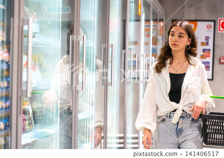 A Young Woman Strolling Among Refrigerator Aisles in a Grocery Store, Exploring Various Food Options and Making Shopping Decisions. 141406517