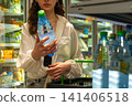 A Person Selecting a Bottle of Water from a Cooler in a Grocery Store Aisle While Shopping for Refreshing Beverages and Other Grocery Items. 141406518