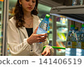 Young Woman Selecting a Bottle of Water from a Refrigerated Display in a Convenience Store, Highlighting Modern Shopping Habits and Healthy Hydration Choices. 141406519