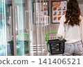 A Woman with a Shopping Basket Walks Through a Grocery Store Aisle, Exploring the Coolers and Reflective Surfaces Amidst Colorful Product Displays. 141406521