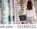 A Young Woman with a Green Shopping Basket Walking through a Modern Supermarket Reflecting on Glass Walls. 141406522