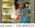 Woman Reaching for Water Bottles in a Convenience Store Fridge Among Other Food Items in a Bright and Organized Shopping Environment. 141406524