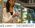 A Young Woman Examining a Bottle of Water in a Store Aisle Amidst Shelves of Various Products While Shopping for Refreshments. 141406525