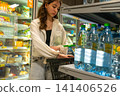 Young Woman Shopping for Bottled Water in a Supermarket Aisle Surrounded by Colorful Snacks and Drinks. 141406526