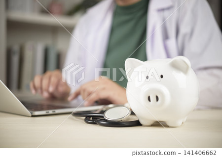 White piggy bank with stethoscope on doctor desk with physician working on laptop in background. Concept of health insurance, medical expenses, healthcare savings, and financial planning. White piggy bank with stethoscope on doctor desk with physician working on laptop in background. Concept of health insurance, medical expenses, healthcare savings, and financial planning. 141406662