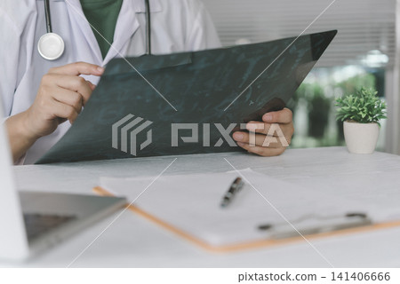 Male doctor holding and examining chest x-ray film at medical office desk. Concept of radiology diagnosis, lung disease analysis, and healthcare checkup. Male doctor holding and examining chest x-ray film at medical office desk. Concept of radiology diagnosis, lung disease analysis, and healthcare checkup. 141406666