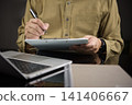 Man hand writing on paper clipboard checklist while working with laptop on office desk. Concept of project management, survey filling, business planning, and inventory checking. 141406667