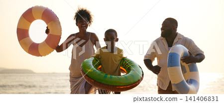 Excited boy holding inflatable ring walking with parents on sunset beach Excited boy holding inflatable ring walking with parents on sunset beach 141406831