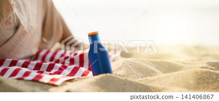Blue sunscreen bottle on sandy beach with red striped towel Blue sunscreen bottle on sandy beach with red striped towel 141406867