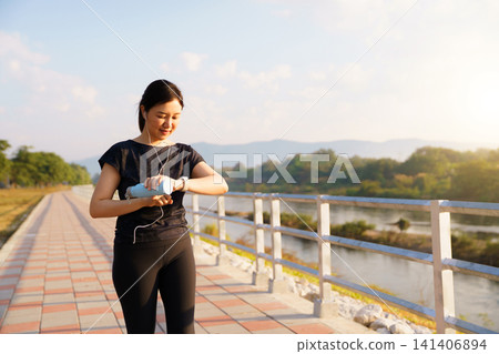 Young Asian woman checking her smartwatch while exercising at a riverside park Young Asian woman checking her smartwatch while exercising at a riverside park 141406894