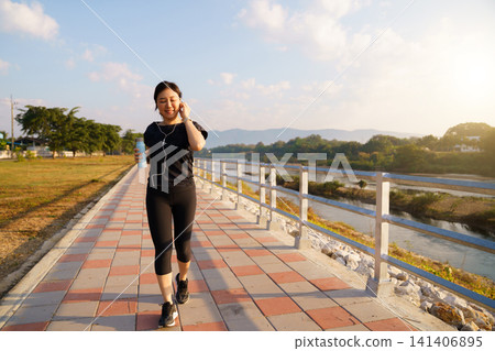 Happy Asian woman jogging along a scenic riverside pathway Happy Asian woman jogging along a scenic riverside pathway 141406895