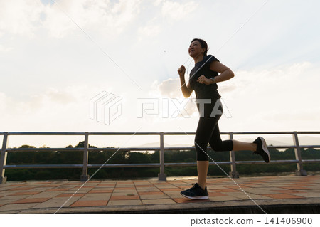 Happy Asian woman jogging along a scenic riverside pathway Happy Asian woman jogging along a scenic riverside pathway 141406900