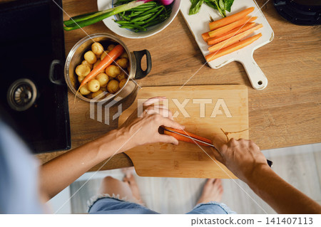Top view of hands slicing fresh carrot lengthwise on wooden chopping board in kitchen Top view of hands slicing fresh carrot lengthwise on wooden chopping board in kitchen 141407113