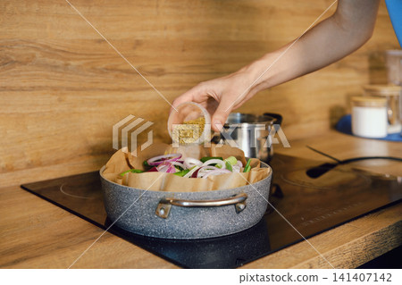Person sprinkles spice and herbs to roasting pot with vegetables in kitchen Person sprinkles spice and herbs to roasting pot with vegetables in kitchen 141407142