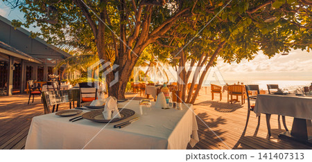 Romantic beach restaurant dining tables under trees at sunset warm lantern light tropical resort terrace. Perfect evening travel hospitality dining outdoors. Luxury honeymoon getaway ambience scenic Romantic beach restaurant dining tables under trees at sunset warm lantern light tropical resort terrace. Perfect evening travel hospitality dining outdoors. Luxury honeymoon getaway ambience scenic 141407313